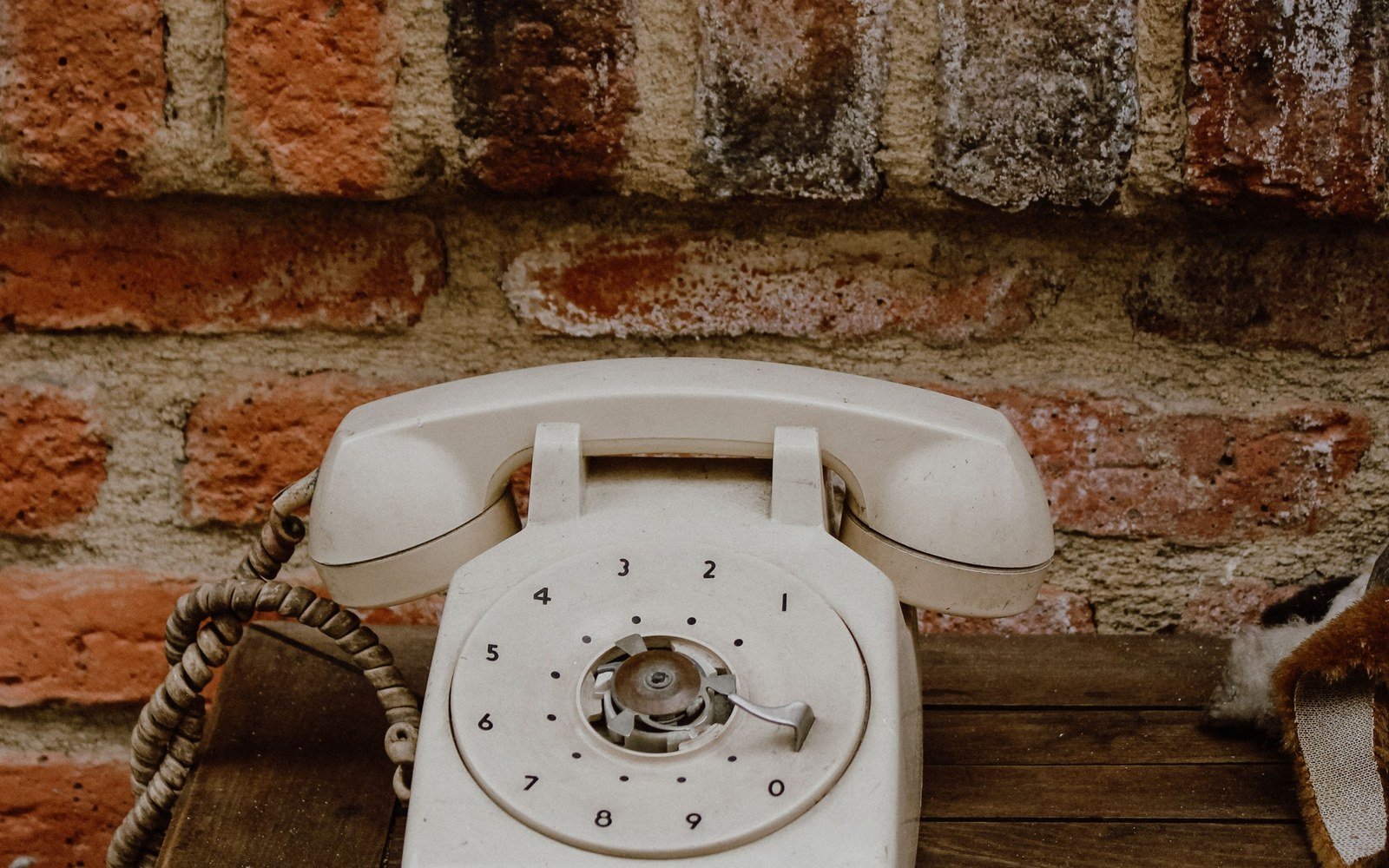 A cream rotary dial telephone with a coiled cord on a wooden table against a brick wall