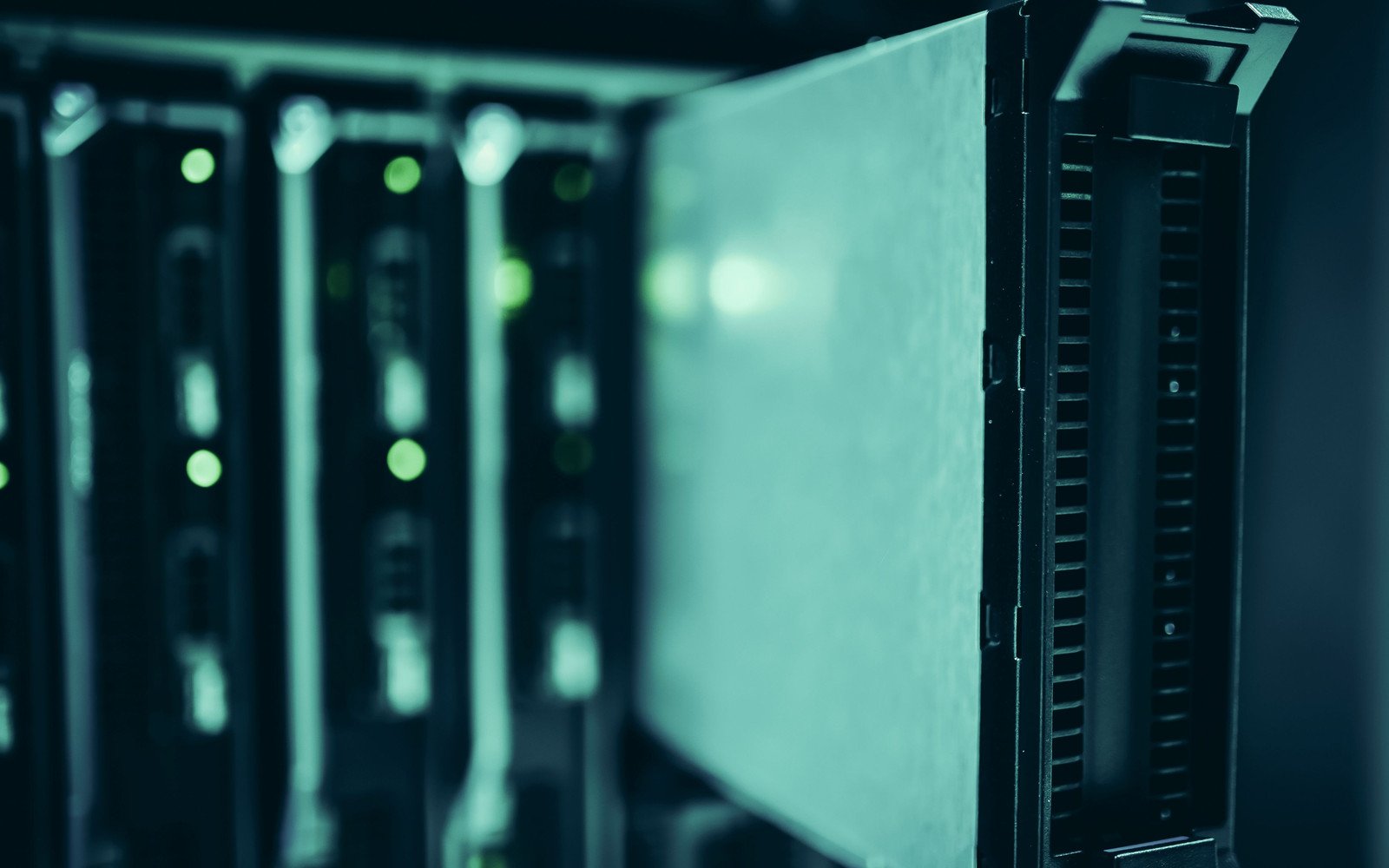 A single server blade partly drawn out of its chassis in close view, lit in green and teal, with a row of further blades and small indicator lights out of focus behind it