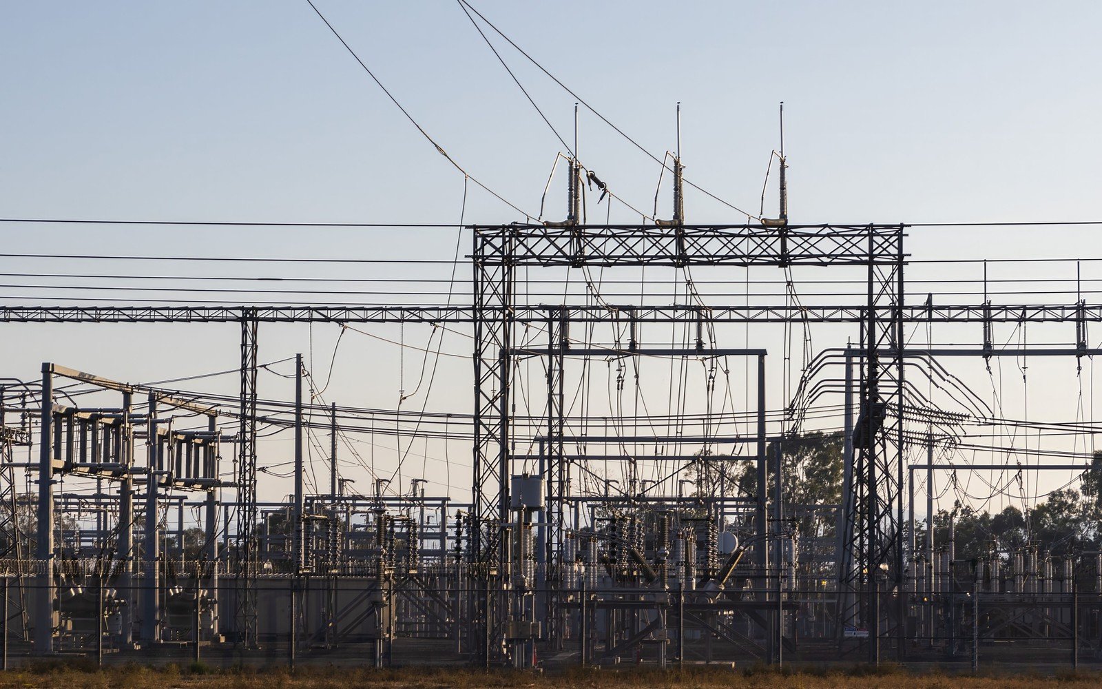 A high voltage substation at dusk, steel gantries carrying insulators and disconnectors, overhead conductors running out of frame in several directions and a low treeline behind