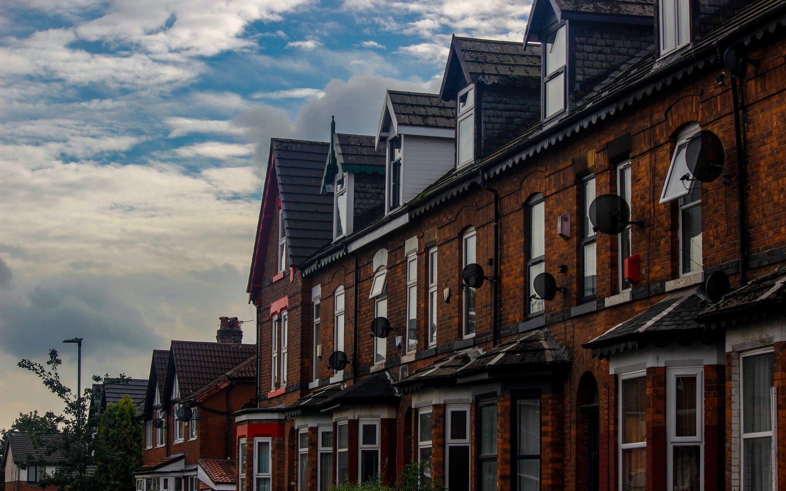 A terrace of red brick houses with bay windows, slate roofs and small dish aerials under a cloudy sky in a Manchester suburb
