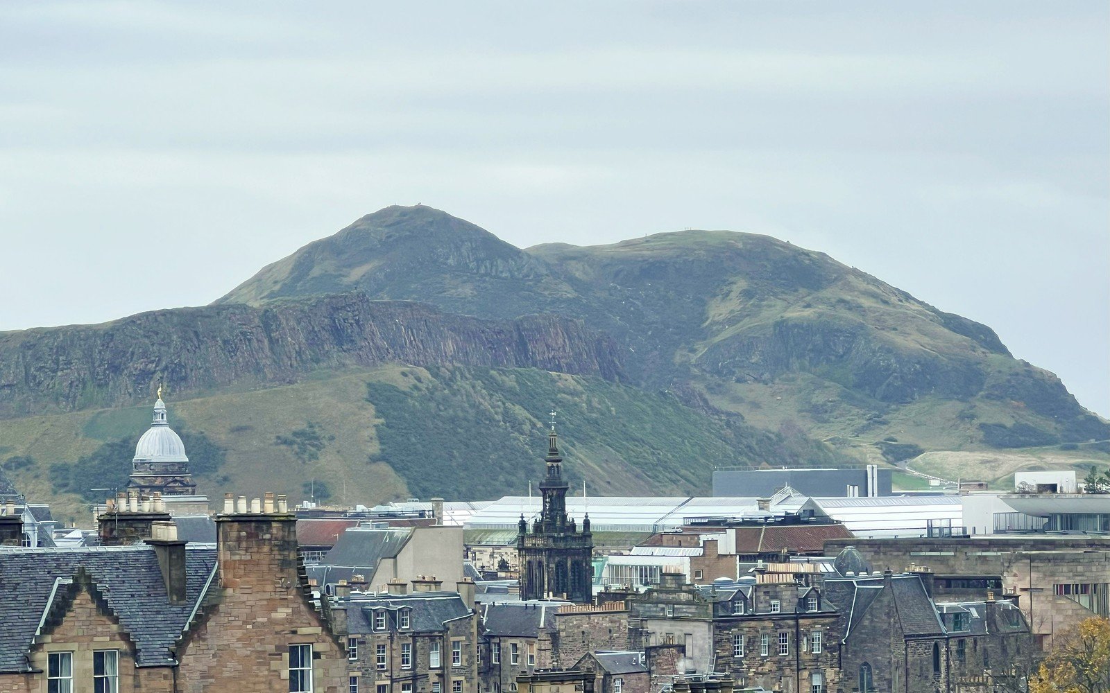 Stone tenement roofs, chimney stacks and church spires below a green hill under a pale sky