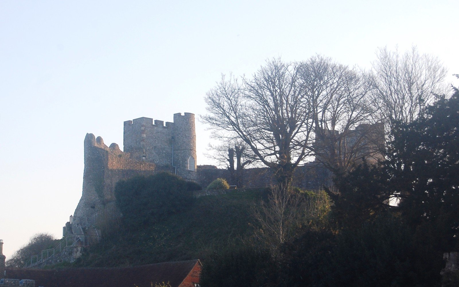 A ruined stone castle keep with two towers standing above trees and a bank on a bright winter day