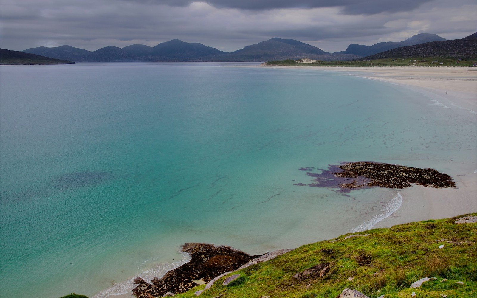 A shallow turquoise bay with a broad pale sand beach curving to a grass bank, and low hills under heavy cloud beyond the water