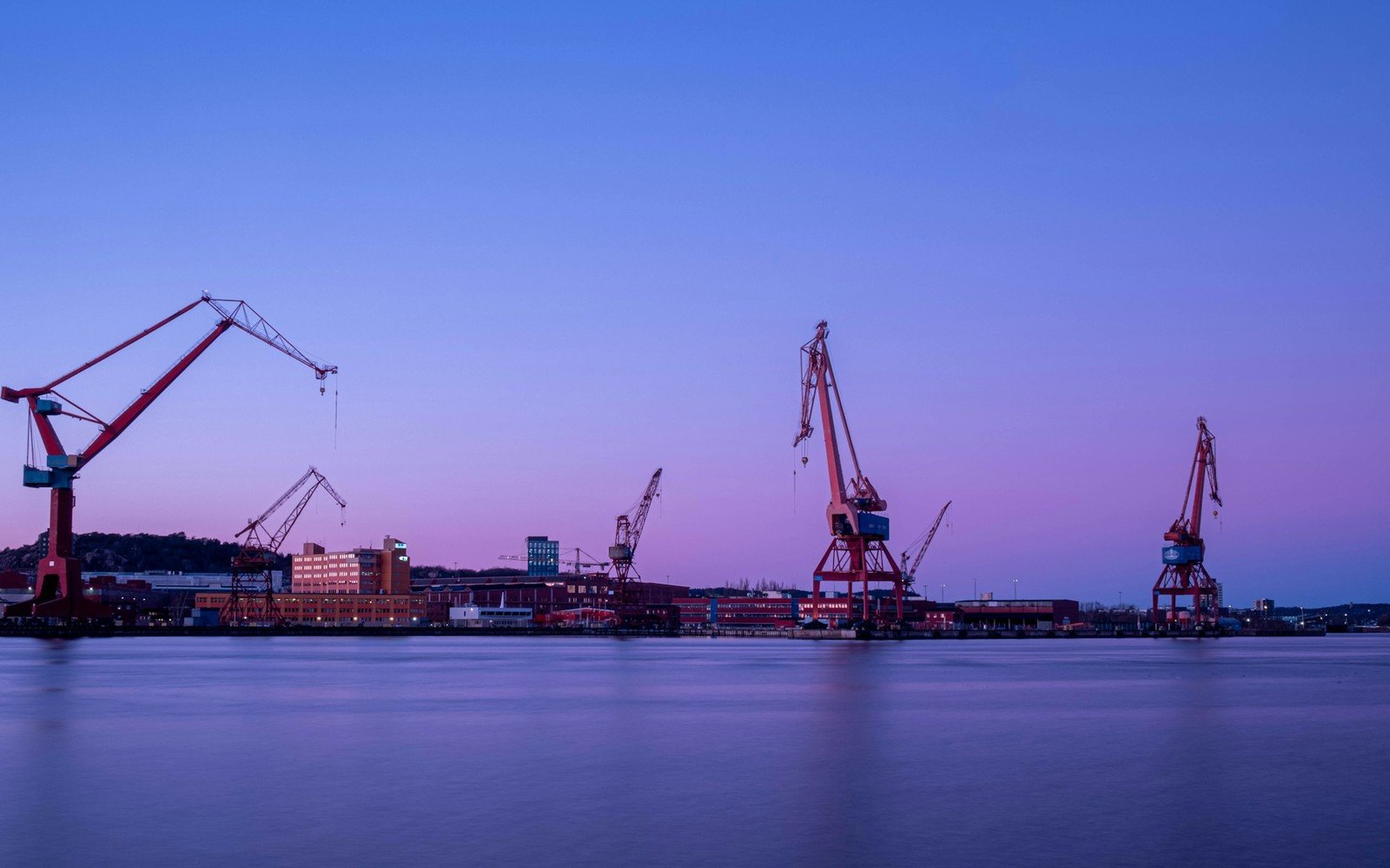 Tall quayside cranes standing over still water at dusk, with low industrial sheds and offices along the far shore under a violet sky