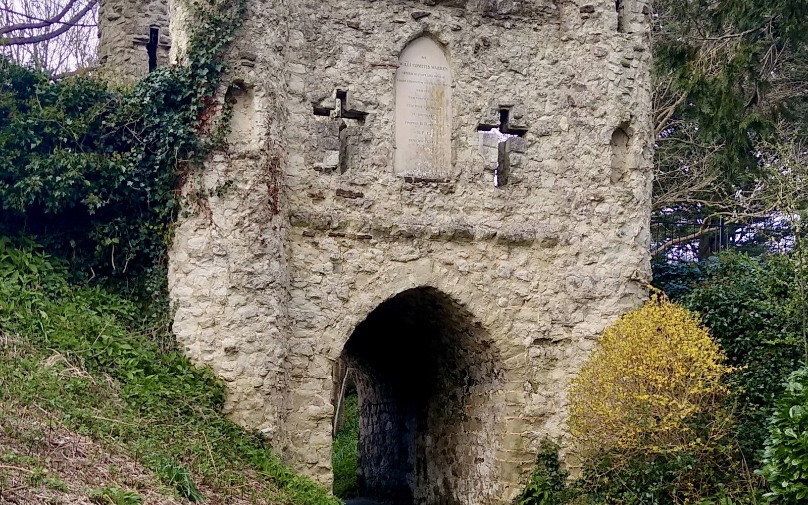 A ruined stone gatehouse with battlements and a pointed arch, overgrown with ivy on a wooded bank
