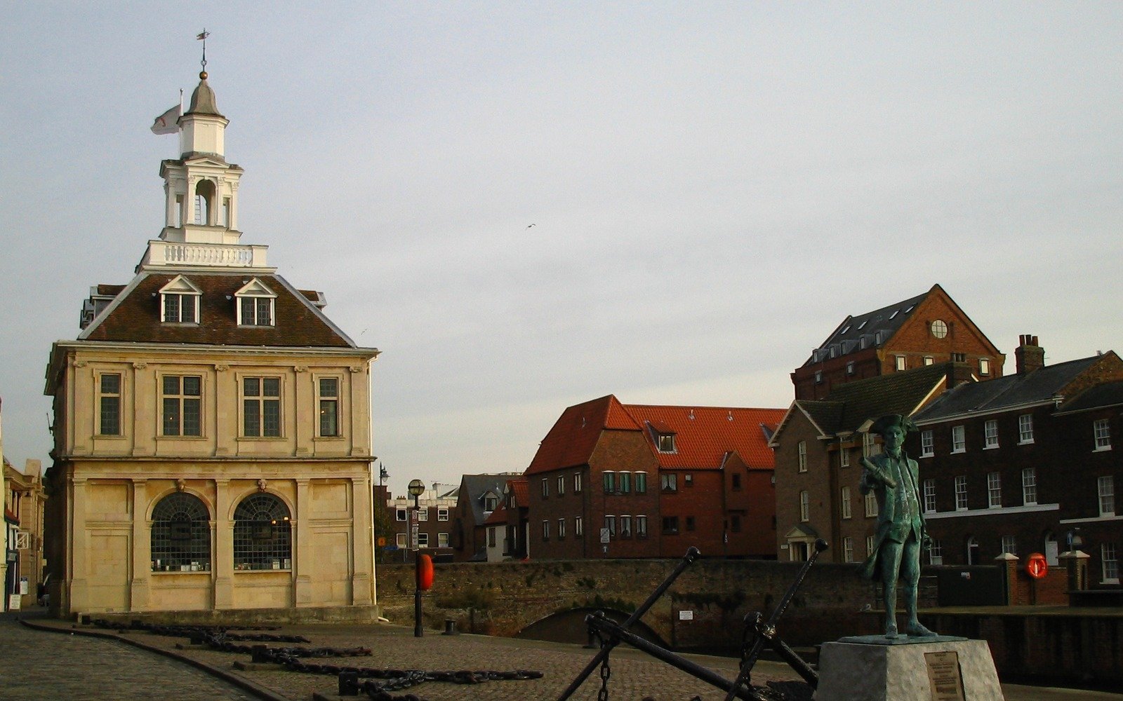 A seventeenth century stone customs building with a lantern cupola and a weather vane, standing on a cobbled quayside beside brick warehouses