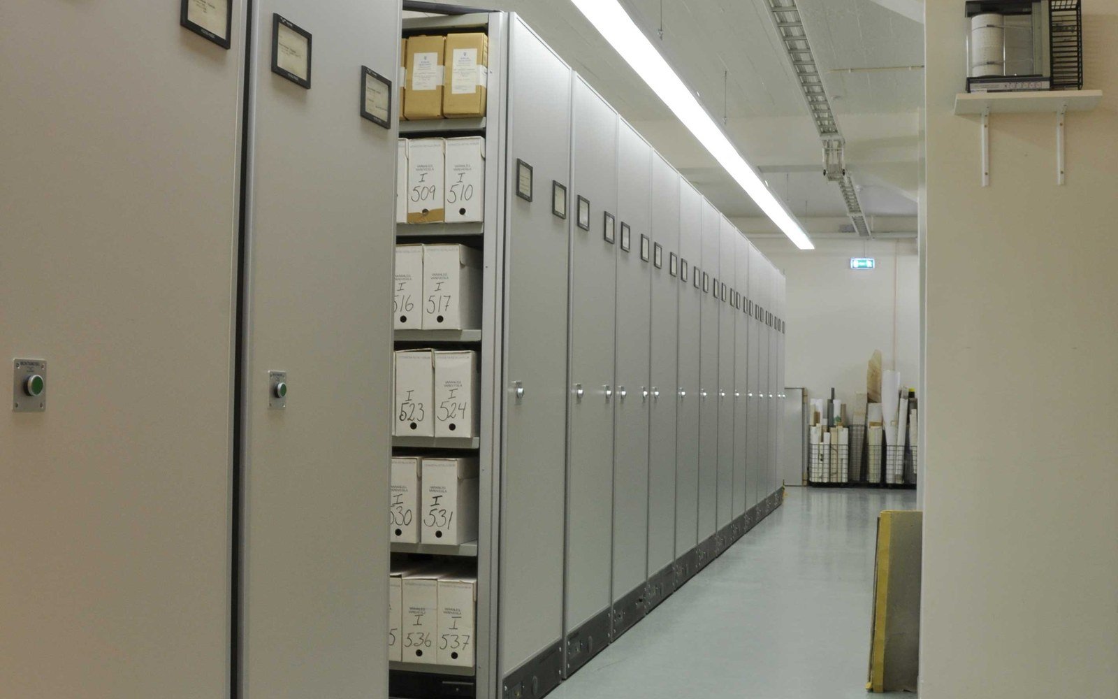 A row of mobile archive shelving units closed together in a corridor, numbered document boxes visible on one open bay