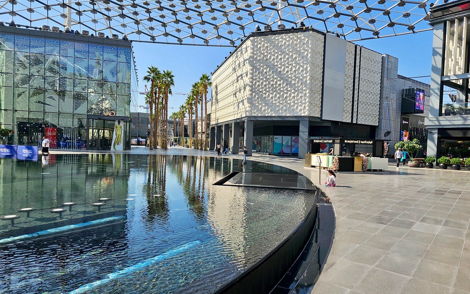 A shallow reflecting pool and paved plaza at City Walk in Dubai, with palm trees, a patterned building facade and a lattice canopy overhead