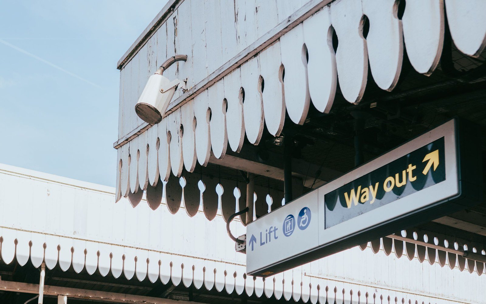 A wayfinding sign reading Lift and Way out hanging beneath a white timber canopy on a London railway platform
