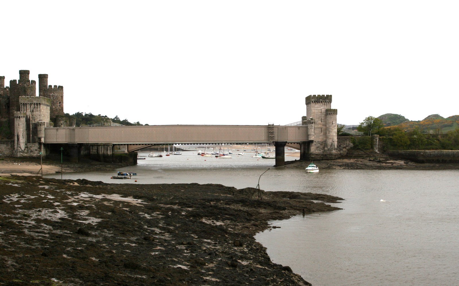 A boxed iron railway bridge crossing a tidal estuary between a castle and a stone tower, with moored boats beyond and mudflats in the foreground
