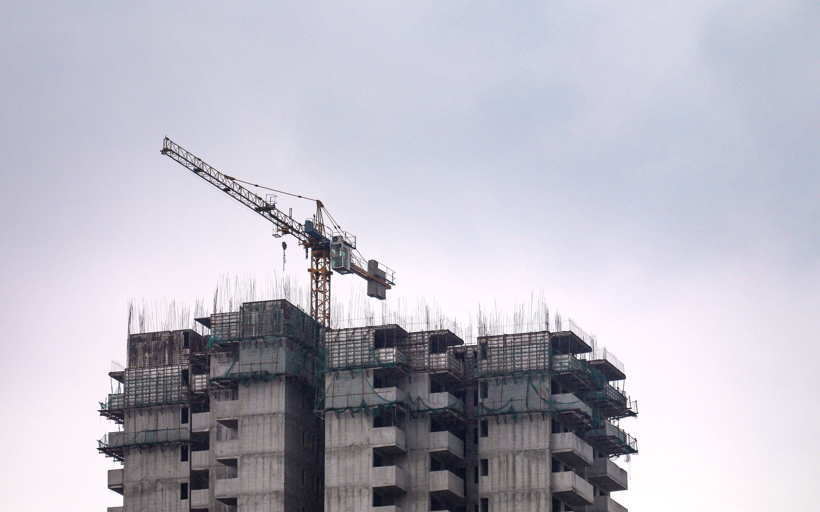The top ten or so storeys of a concrete residential frame under construction, starter bars projecting from the roof slab, green netting across the open edges and a tower crane standing over it against a grey sky