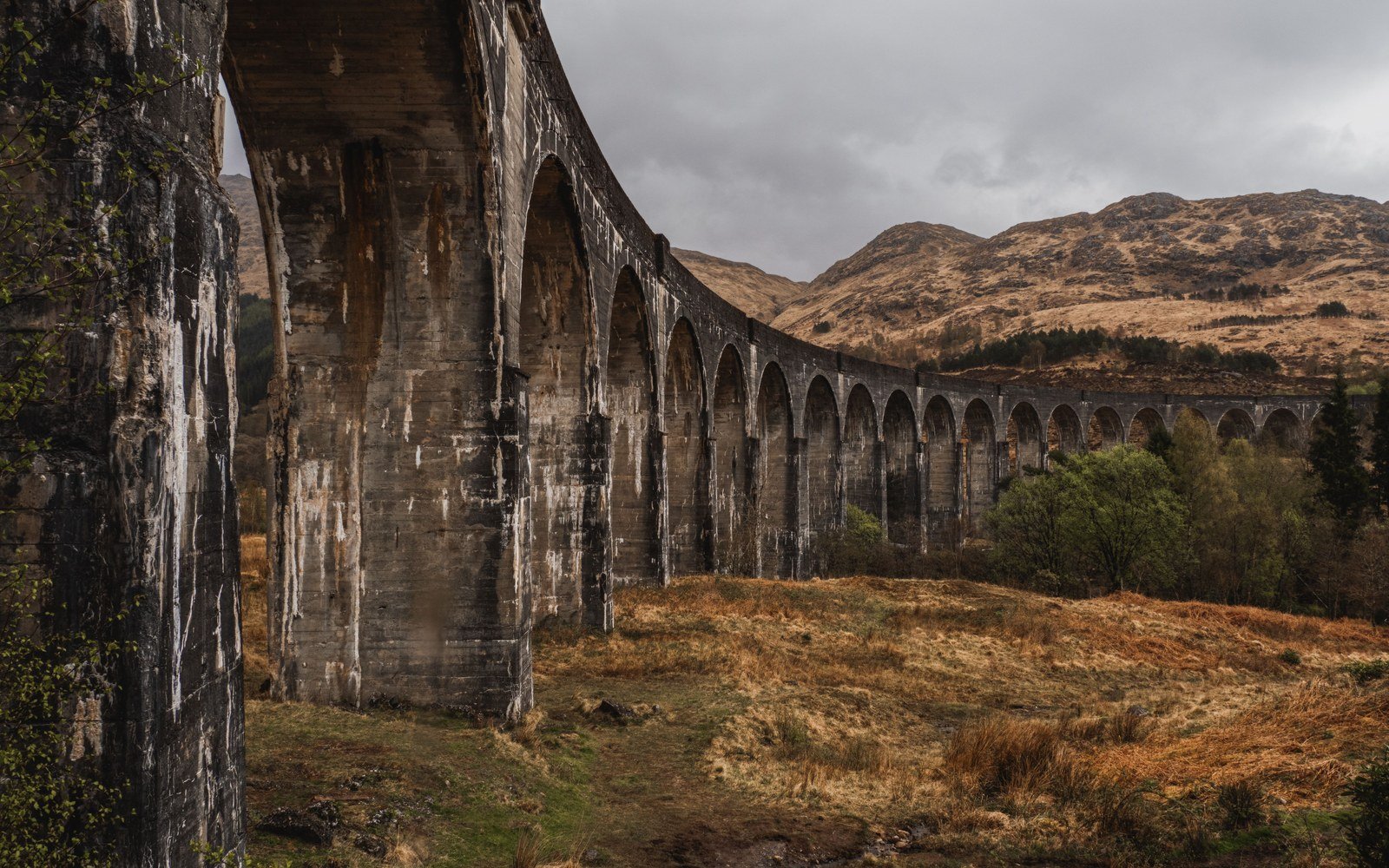 A tall concrete viaduct of curving arches carrying a railway across a glen, bracken covered hillsides rising behind it under heavy cloud