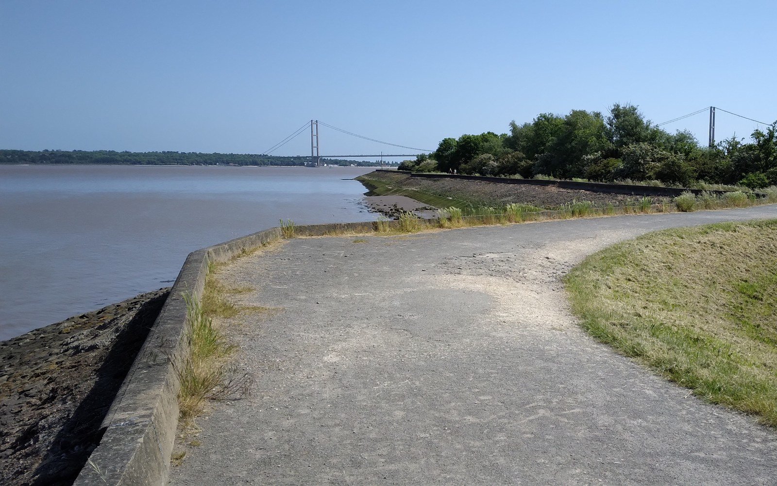 A suspension bridge crossing a wide tidal estuary, seen along a surfaced shoreline path from the southern bank