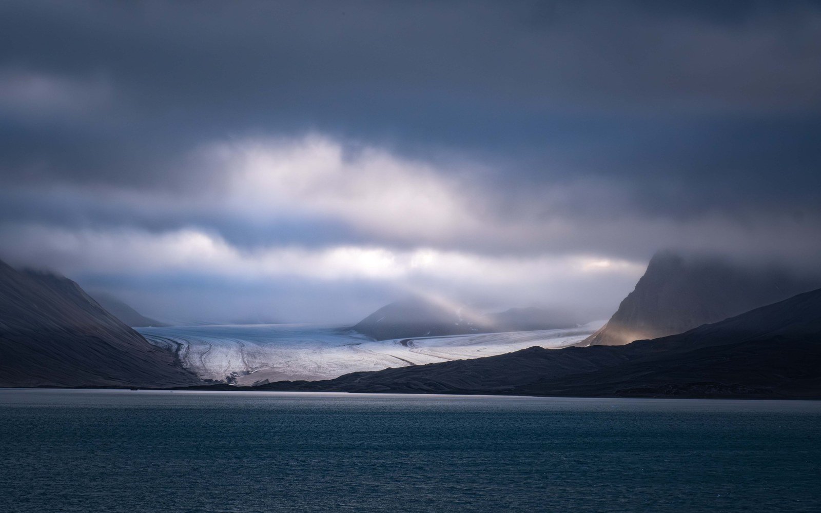 A glacier front between dark mountains under heavy cloud above still water in Svalbard