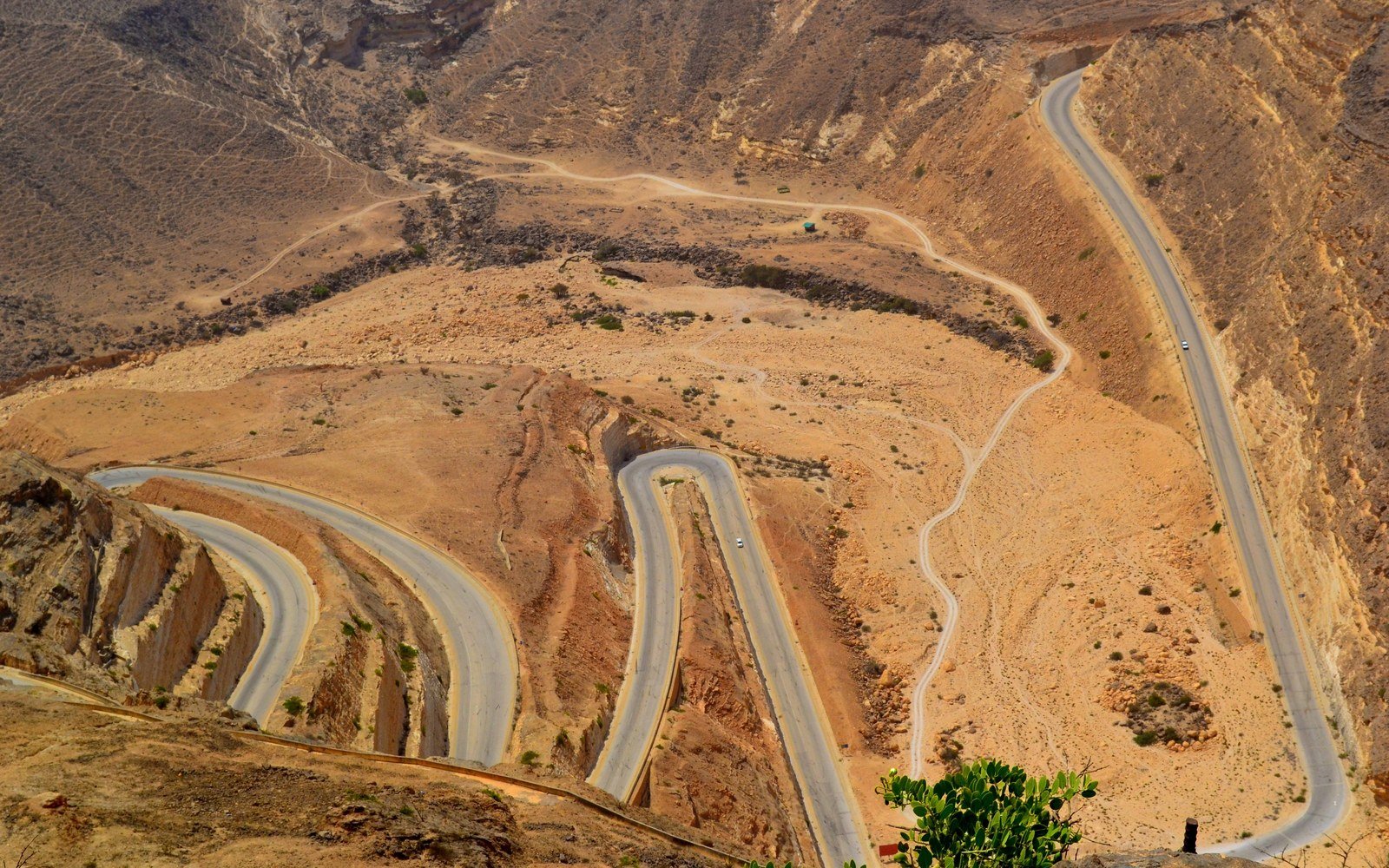 A single carriageway road climbing in tight hairpin bends across bare rock and gravel slopes in the Dhofar mountains of Oman, a second road running along the valley floor, no vehicles or people visible