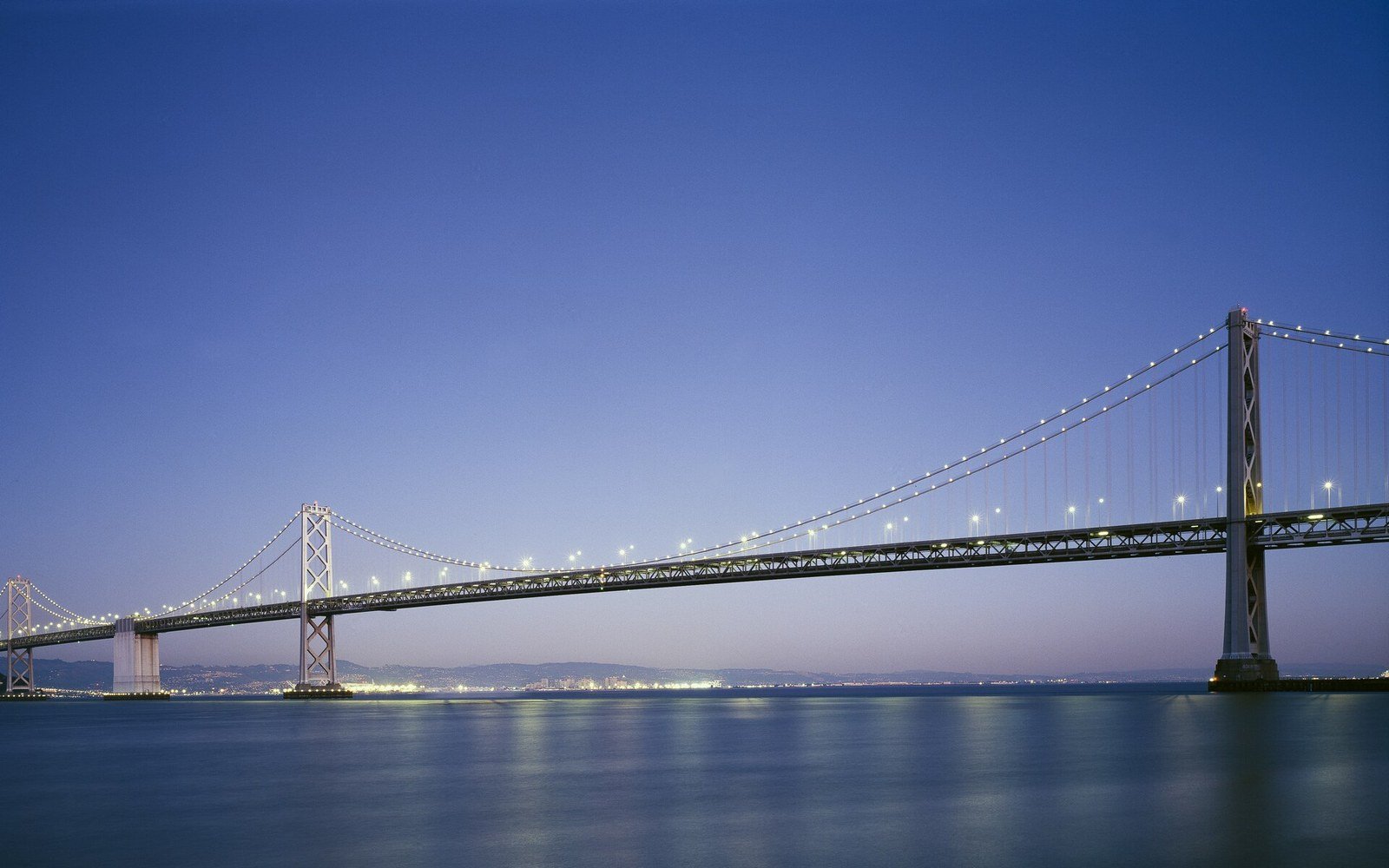 The San Francisco to Oakland Bay Bridge at dawn, its lights still on, with the bay flat and the city lit low on the far shore