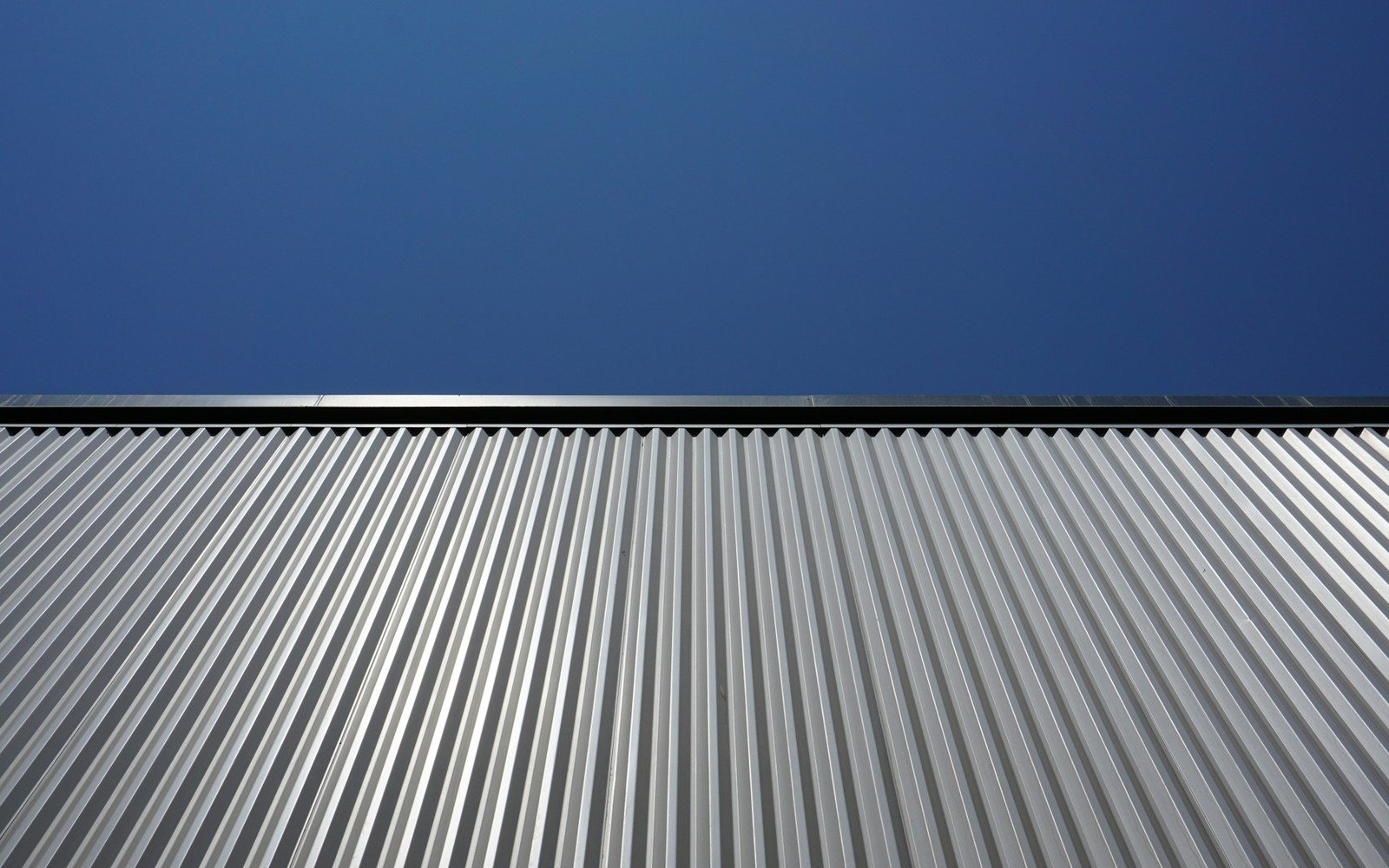 A close view along a pitched roof of corrugated metal sheeting, the ribs running away from the camera under a deep blue sky