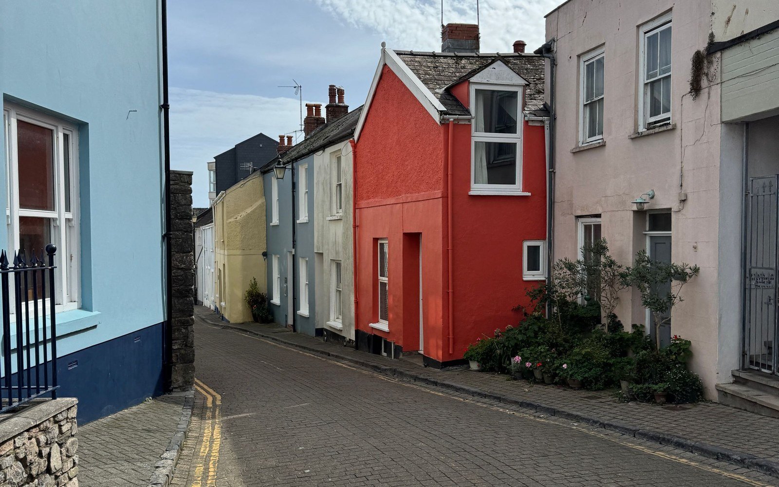 A narrow street of rendered houses painted red, cream, blue and grey in a Welsh town, with a low stone wall and railings in the foreground