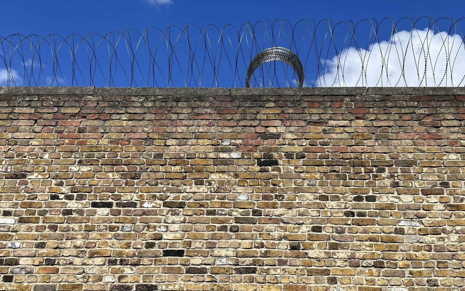 A weathered brick boundary wall topped with coiled razor wire against a clear blue sky