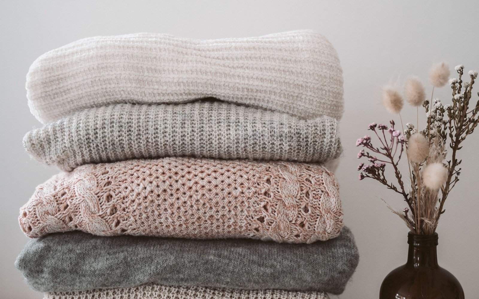 A neat stack of six folded knitted jumpers in cream, grey and pink on a pale painted surface, with a small vase of dried flowers beside it
