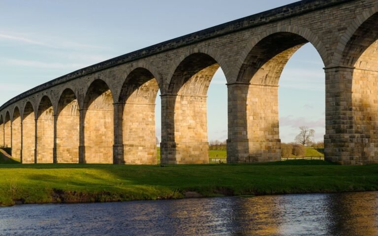 The stone arches of a long curving railway viaduct in low sunlight, with a river in the foreground