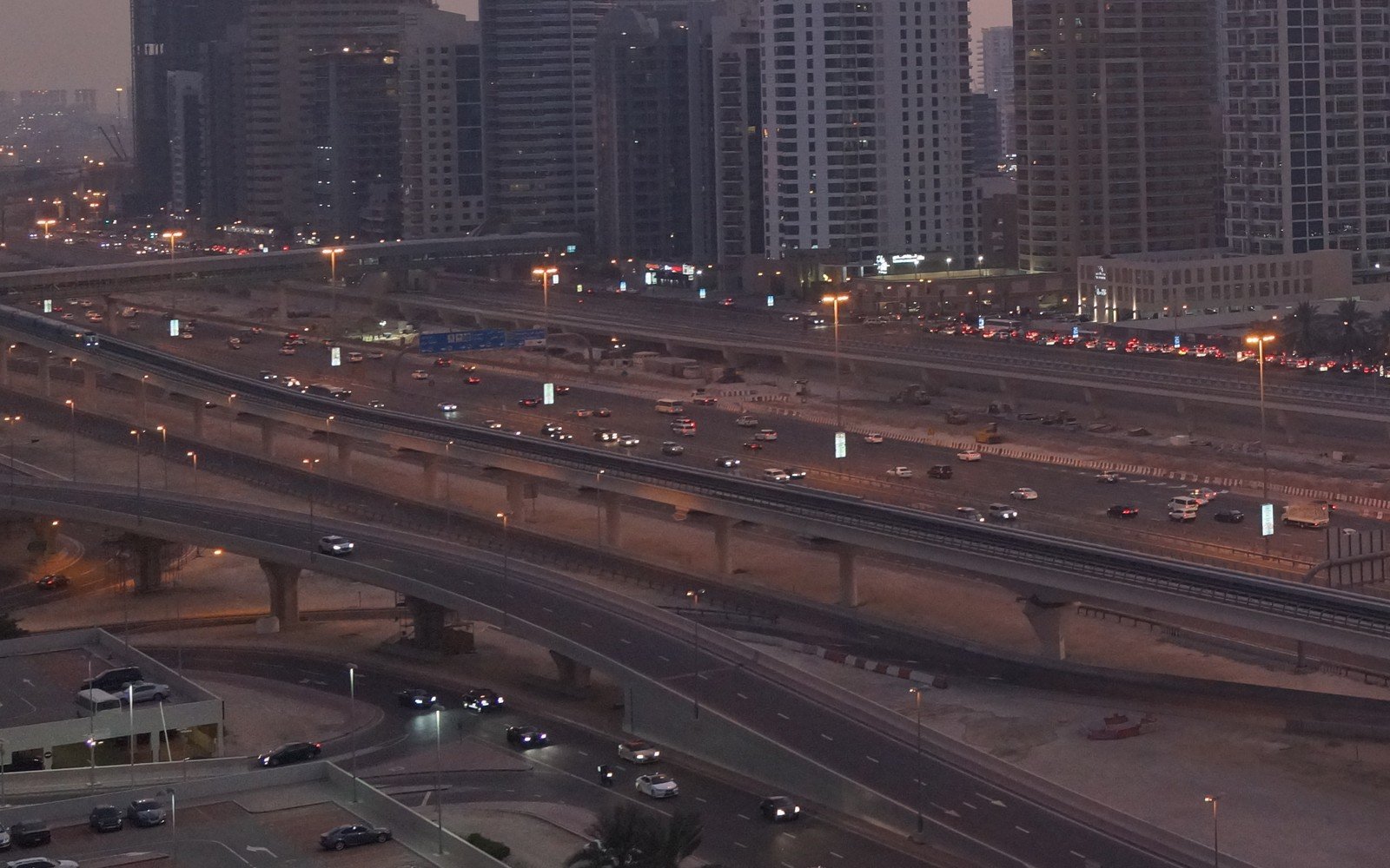 A multi-lane road corridor in Dubai at dusk, with an elevated flyover and a raised metro viaduct running between residential towers