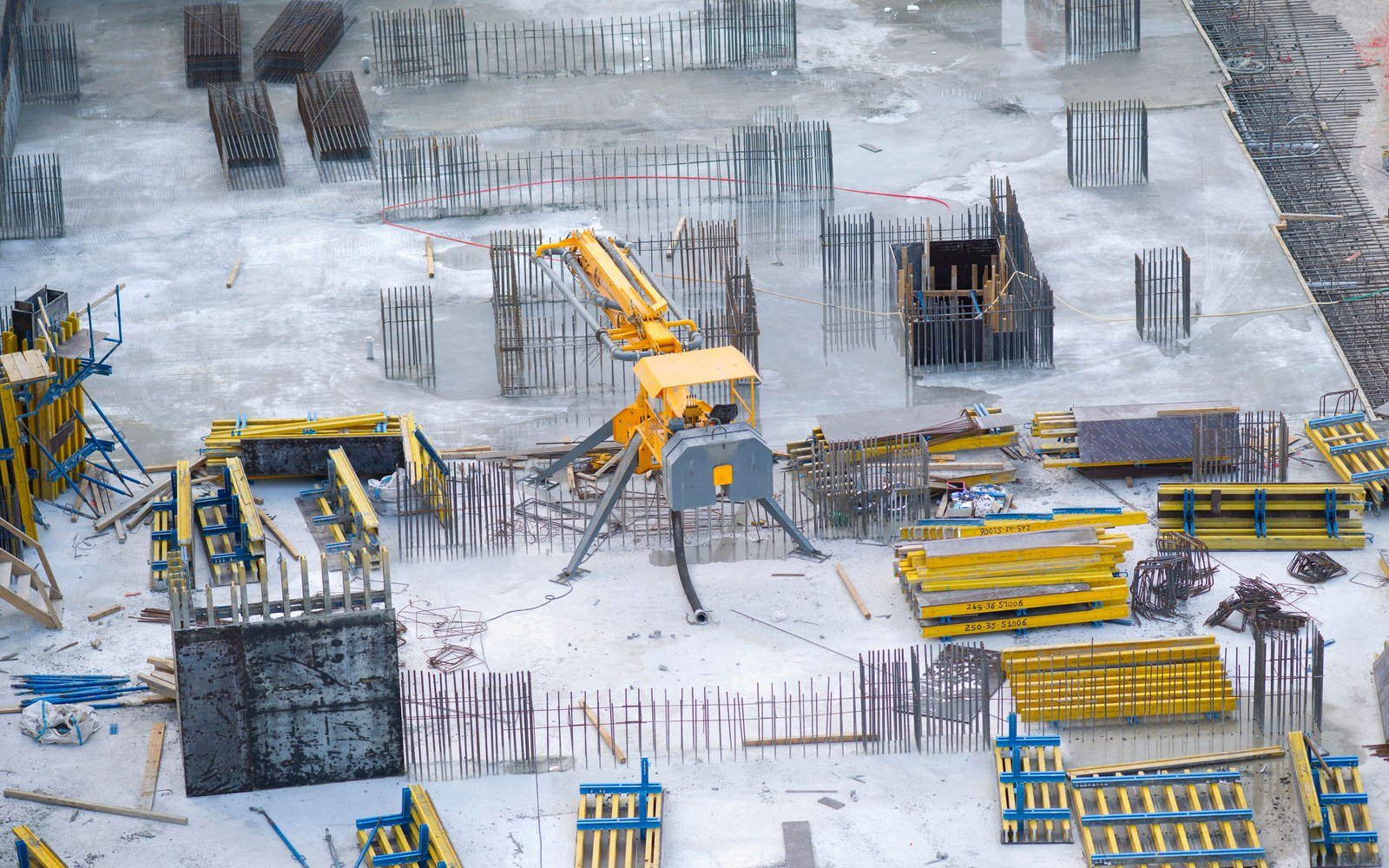 An overhead view of a reinforced concrete slab under construction, with column starter bars projecting through the deck, yellow and blue formwork panels stacked flat, loose reinforcement and a concrete placing boom standing on its outriggers