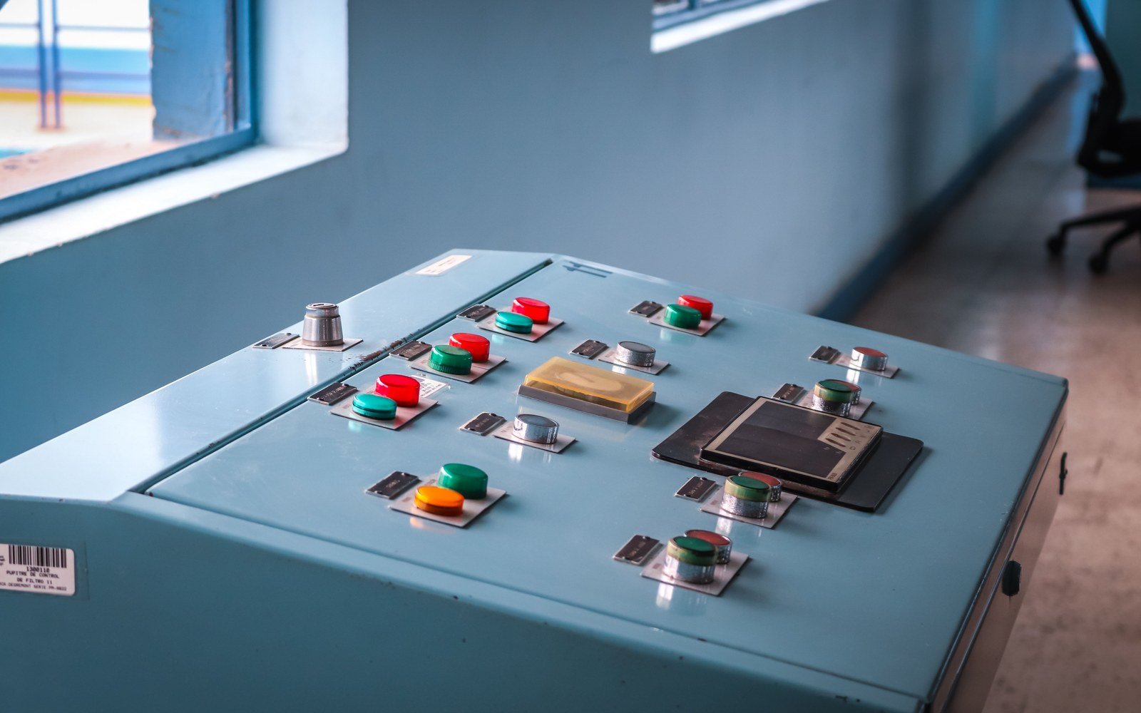 A pale blue industrial control desk photographed close up, rows of red, green and orange push buttons on steel plates, a hinged instrument cover and a window looking out to a steel walkway