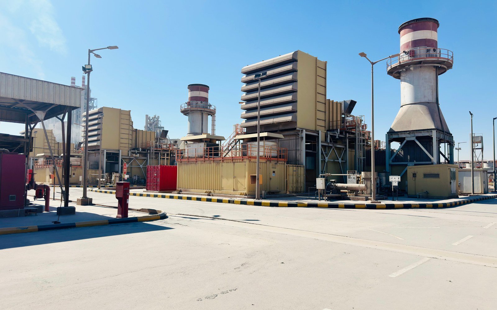 Gas turbine enclosures, filter houses and two red and white banded exhaust stacks at an industrial plant in Saudi Arabia, seen across an empty concrete apron under a clear blue sky