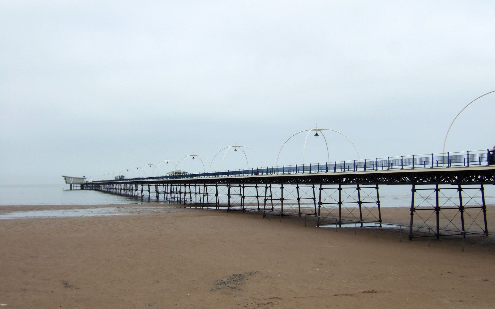 Southport Pier stretching out across wet sand towards the sea under a grey sky, its iron legs and arched lamp standards in a long line