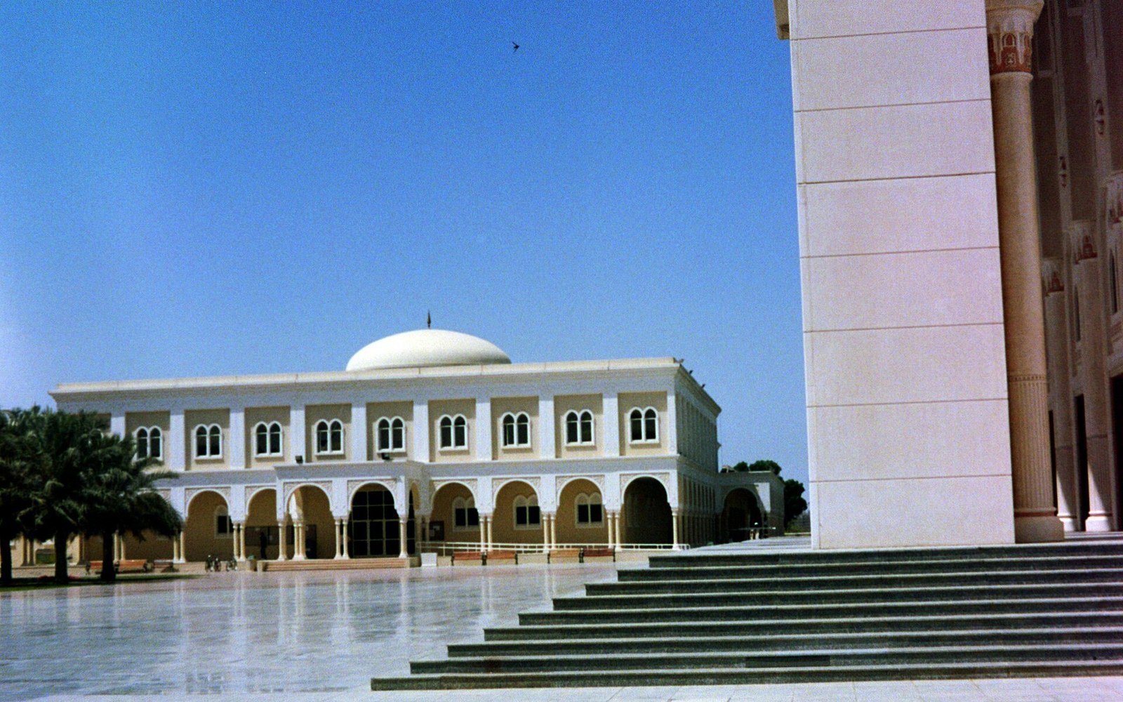 A cream coloured university building with arched colonnade and a white dome behind a paved forecourt and steps in Sharjah, under a clear sky