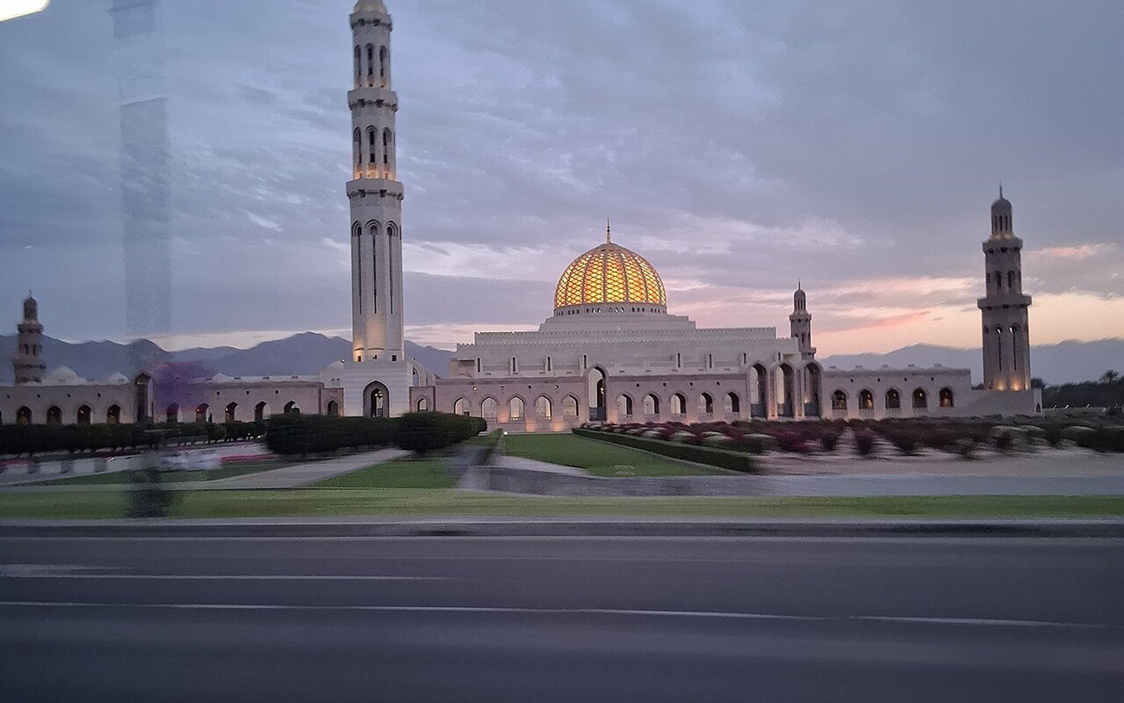 Sultan Qaboos Grand Mosque with its gold dome and minarets seen across a road and lawns at dusk