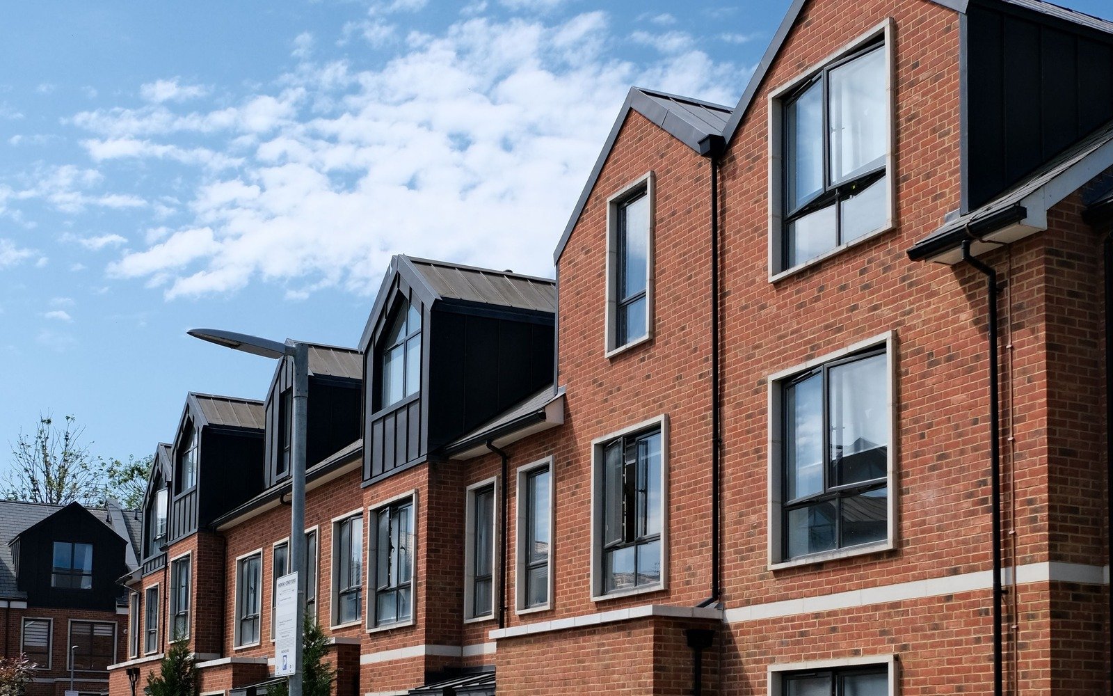 A row of newly built three storey brick houses with gabled roofs and tall white framed window units, photographed from the pavement against a blue sky