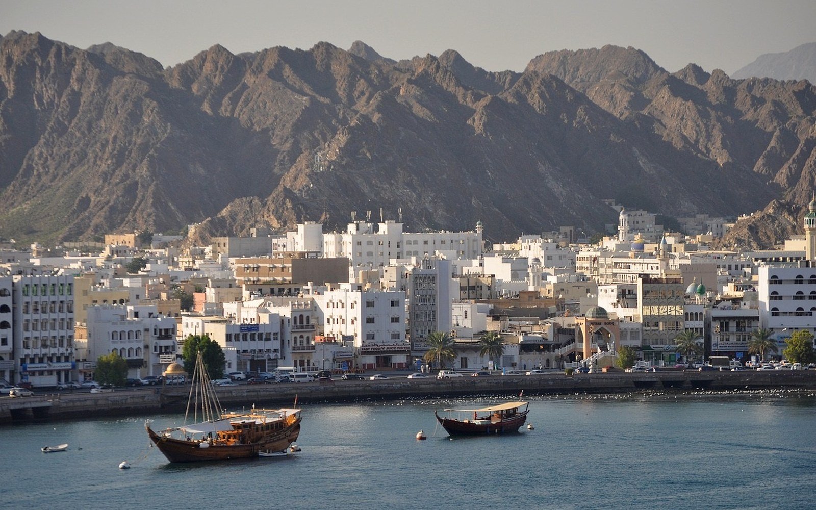 The Muscat waterfront at low sun, white low rise buildings along a corniche below the bare rock ridge of the Hajar mountains, with two wooden dhows moored on flat water in the foreground