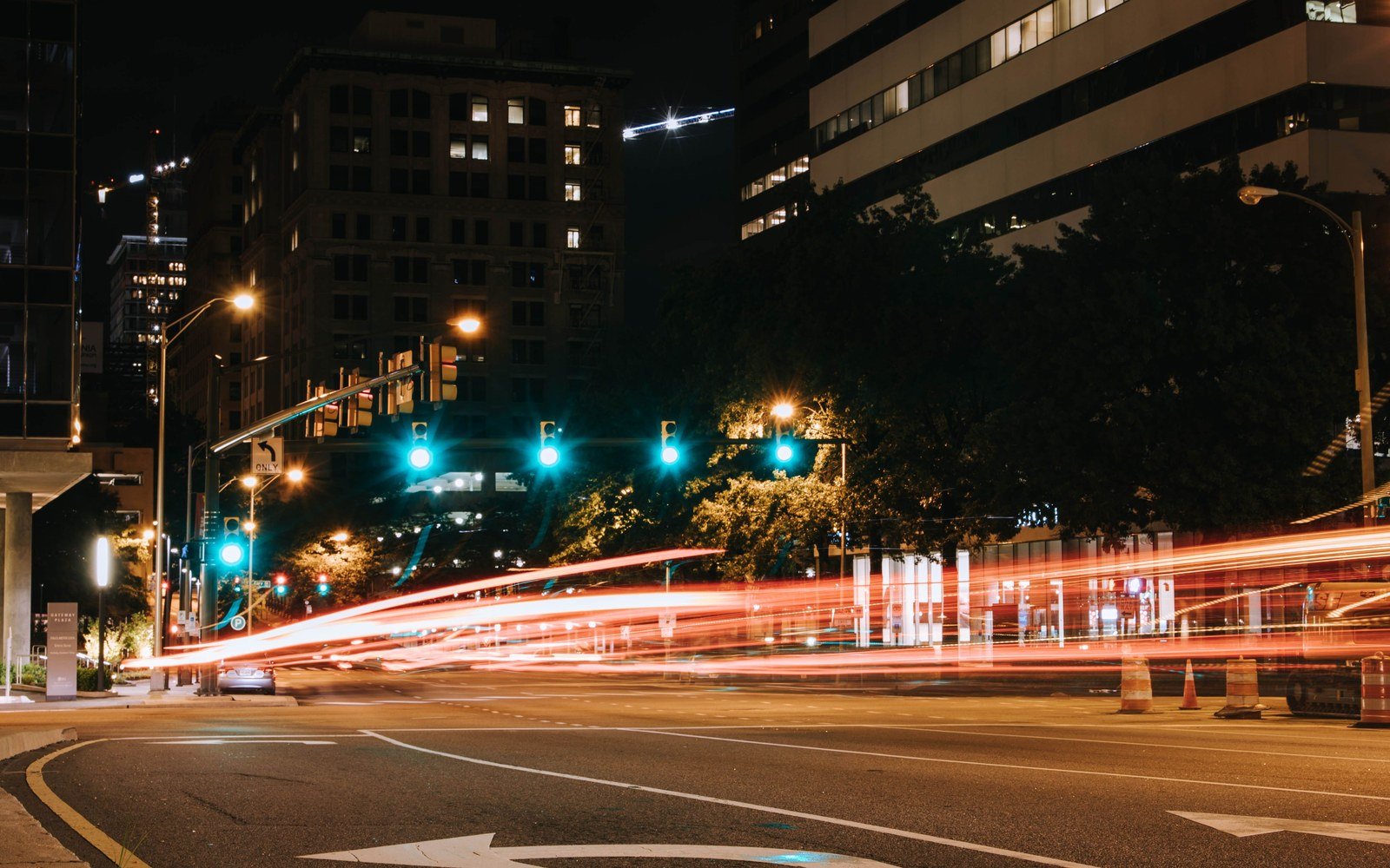 A downtown city street at night with green traffic signals and the red light trails of passing cars, tall buildings on either side
