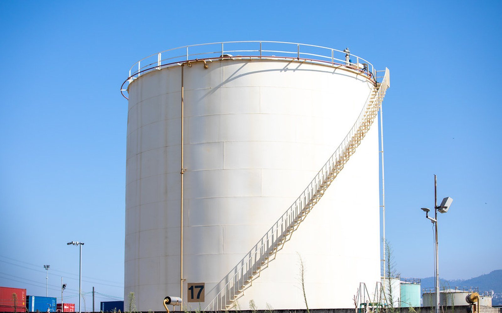 A large white cylindrical storage tank with a spiral stair to its roof, standing against a clear blue sky