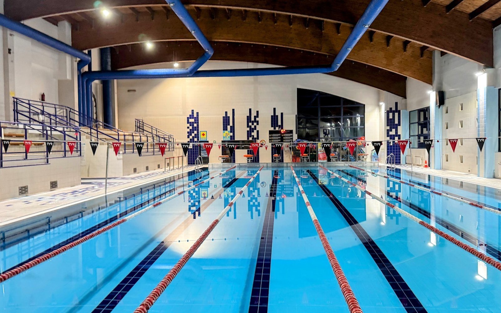 An empty indoor swimming pool with marked lanes and blue water under a timber and steel roof