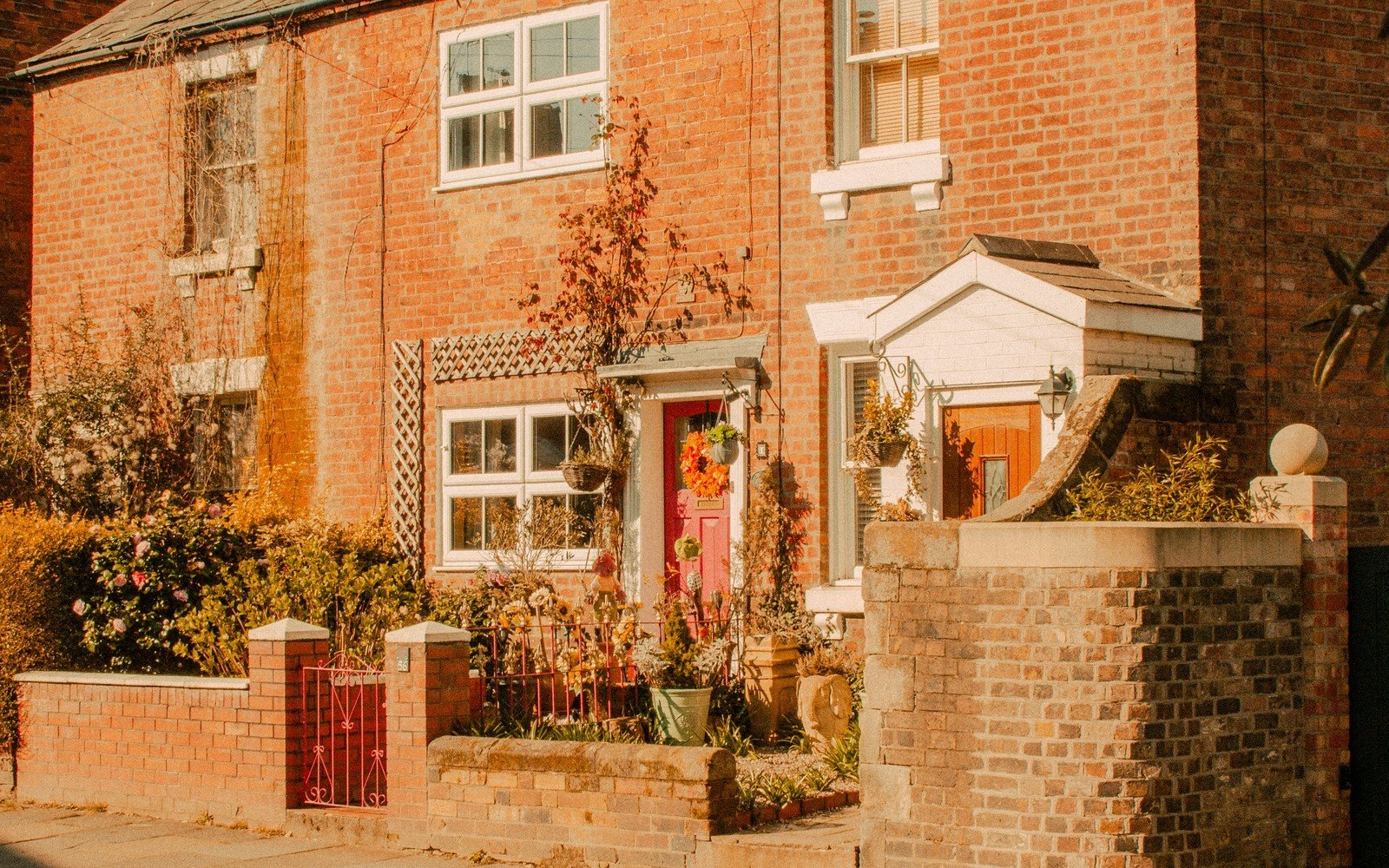 The brick front of a house with a red door, a white porch and a planted front garden behind a low brick wall in sunshine