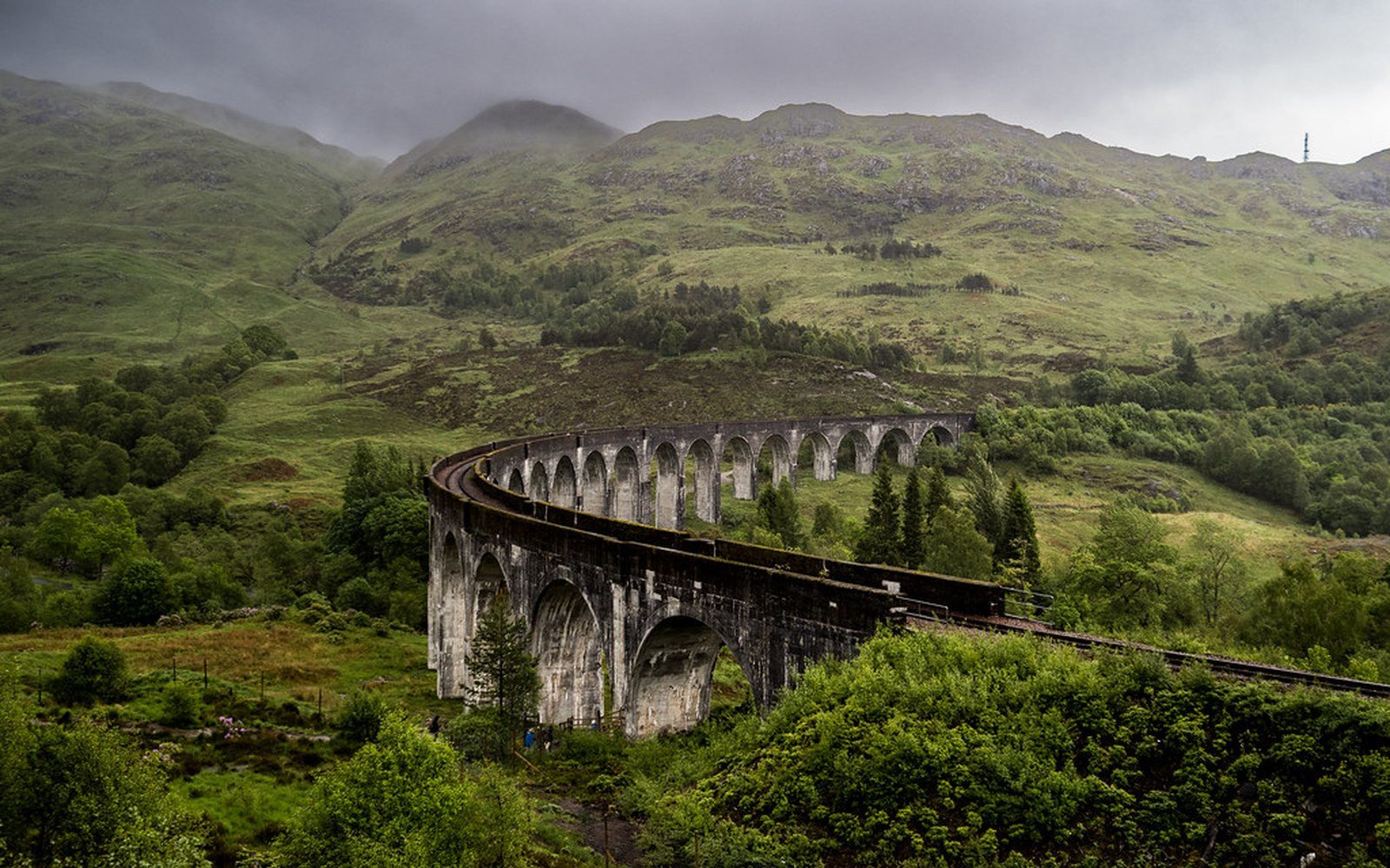 The curved concrete arches of Glenfinnan Viaduct carrying the railway across a green glen, with cloud on the hills behind