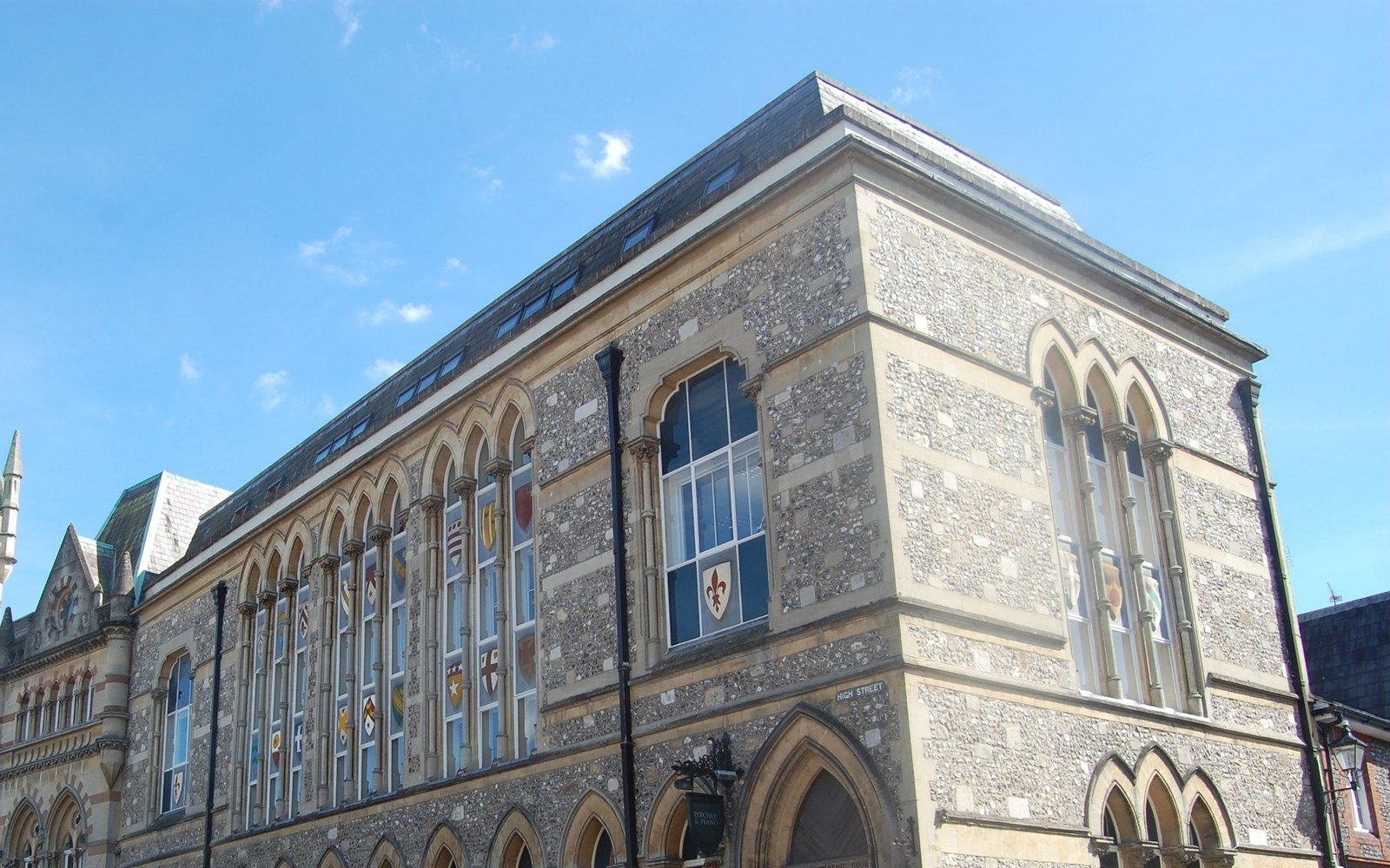 The flank of a Victorian Gothic municipal building in flint and stone, with tall arched windows and a steep slate roof against a clear sky