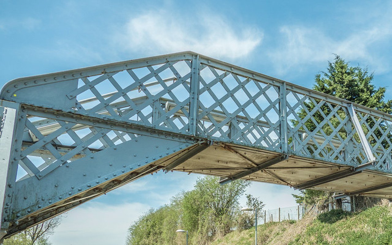 A riveted steel lattice footbridge spanning a railway line against a blue sky, with trees on the embankment below