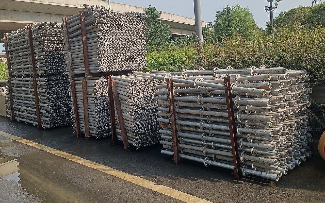 Bundles of galvanised steel tubes in rusted steel frames stacked beside a wet road