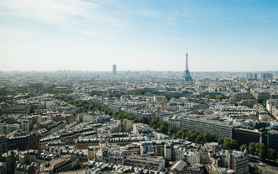 The rooftops of western Paris seen from a tall building, with the Arc de Triomphe and the Eiffel Tower under a hazy blue sky