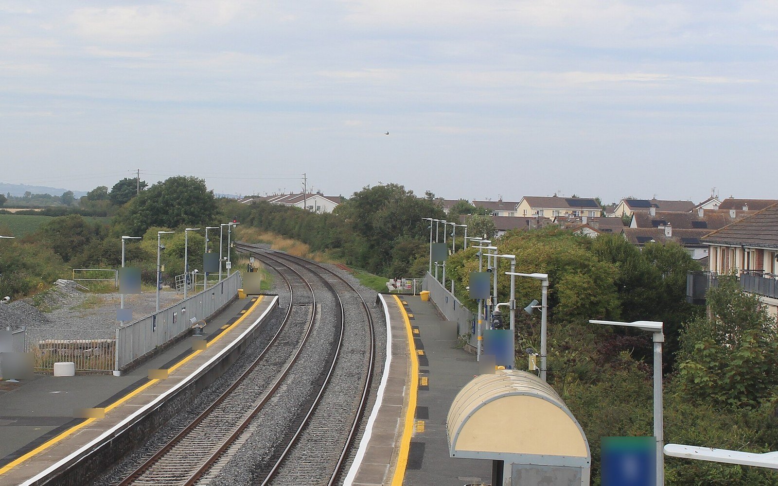 A curving double railway track between two platforms with yellow edge lines, lamp posts and a canopy, with houses and trees beyond under a grey sky