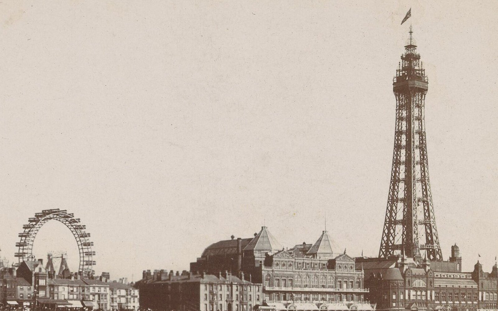 An early sepia photograph of a tall lattice tower and large Victorian buildings along a seafront, with a big wheel to one side