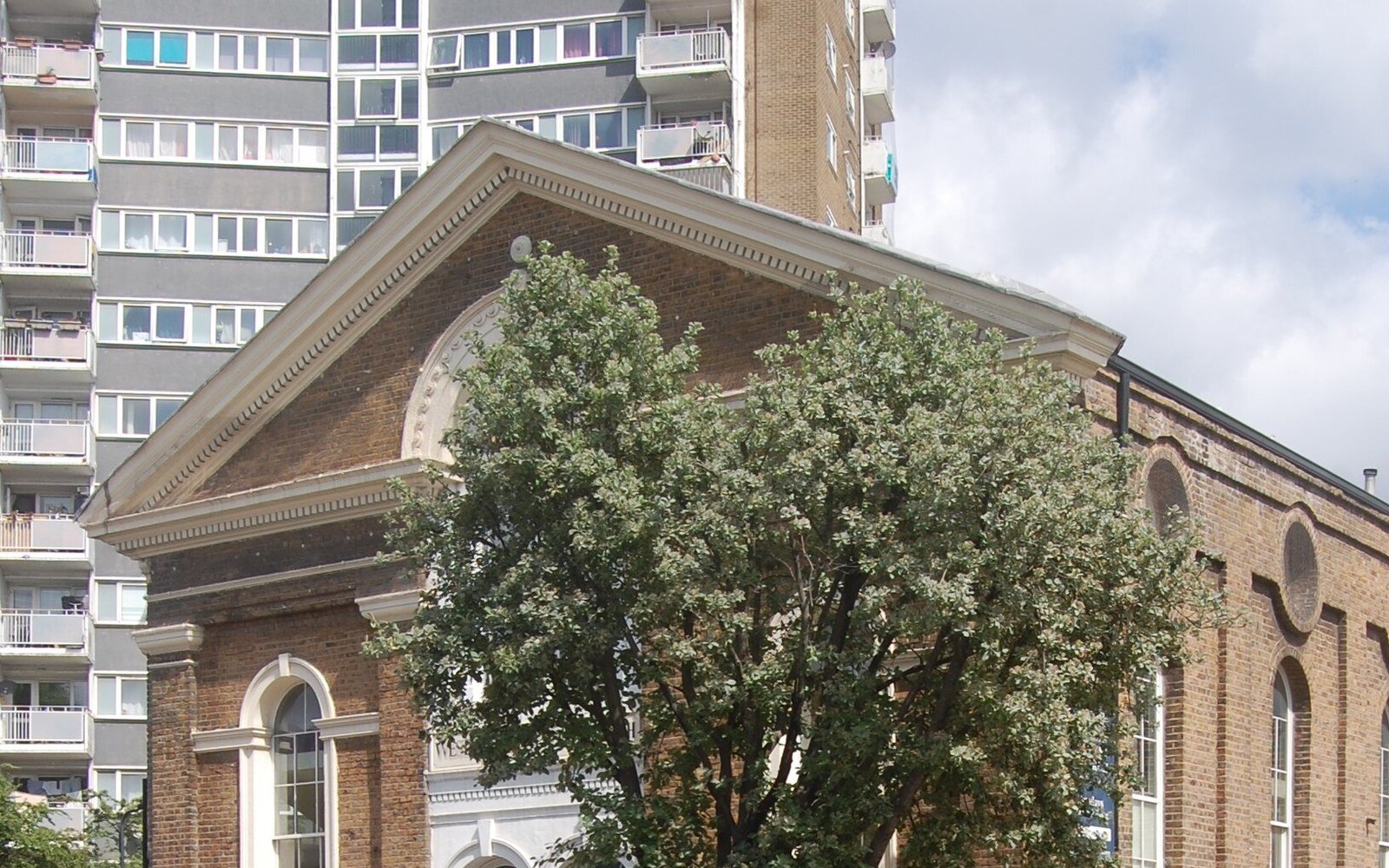 A brick chapel with a classical pediment and arched windows partly hidden by a tree, in front of a tall residential tower block with balconies