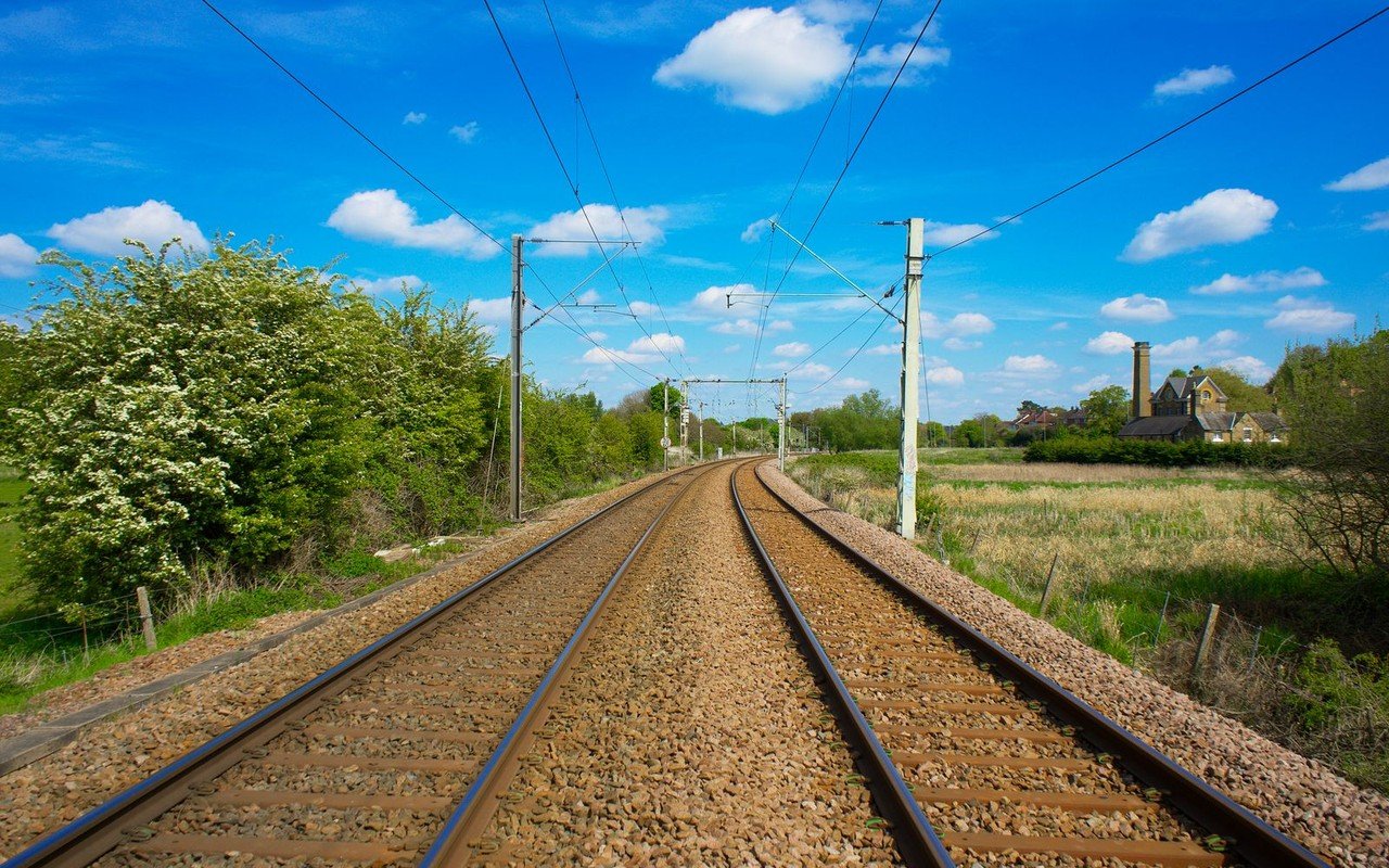 Twin railway tracks running straight into the distance between hedgerows and a meadow, with overhead line equipment on masts under a blue sky with white clouds