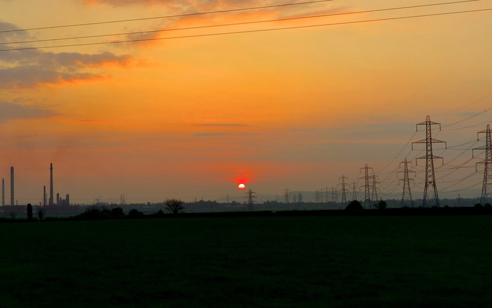 An orange sky at sunrise over flat dark fields, with a row of electricity pylons, overhead lines and distant chimneys on the skyline