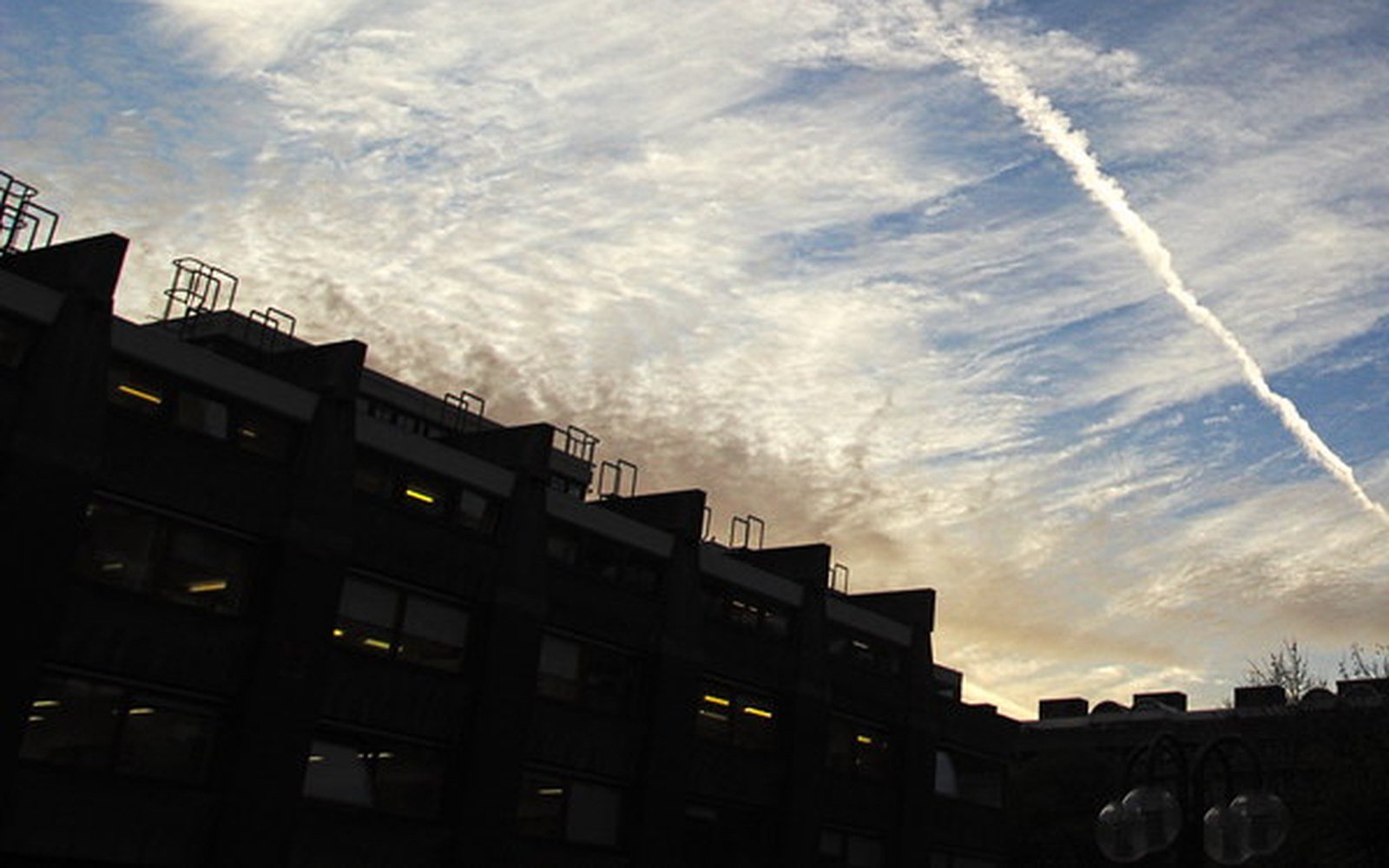 The dark silhouette of a long stepped concrete residential block against a sky streaked with white cloud and a vapour trail at sunset
