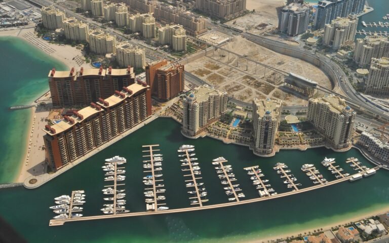 An aerial view of a curved marina with rows of moored boats, ringed by tall apartment blocks, sandy beaches and turquoise water